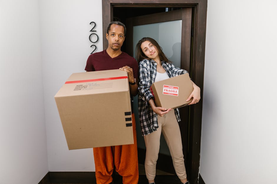 A man and a woman are indoors, preparing for a home relocation by carrying packing and moving supplies. The man is holding a small cardboard box labeled fragile with one arm, while the woman is holdin
