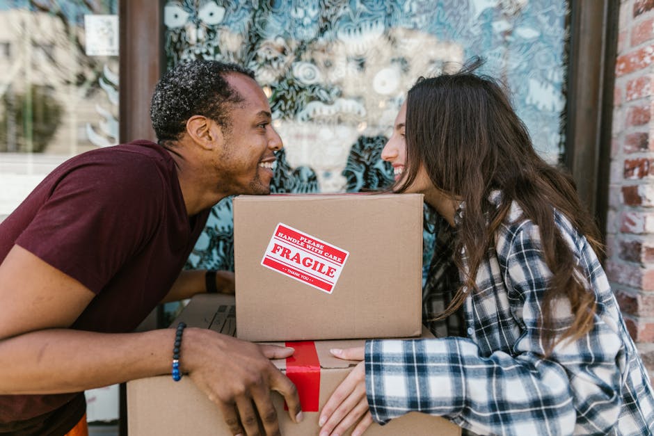 A man and a woman are outdoors, facing each other with their foreheads touching, both smiling. The man, with curly dark hair and wearing a maroon T-shirt, is holding a cardboard box with red and white 'Fragile' packaging and handling tape. The woman, with long brown hair and dressed in a blue and white checked shirt, is supporting the box with her hands. They are standing in front of a window with decorative frosted glass and a brick wall, suggesting a home environment. The scene captures a moment during a home relocation process involving packing and furniture transport, with the couple demonstrating care and cooperation in handling boxes for a move, facilitated by a professional removals service like Man With a Van Brockley.