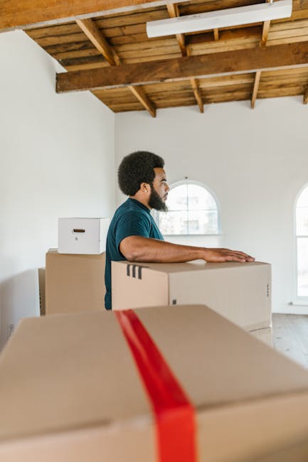 A man with dark curly hair and a beard, wearing a navy blue t-shirt, is seen inside a bright, spacious room with white walls and a wooden ceiling with exposed beams. He is standing behind several moving boxes, which are made of cardboard and sealed with packing tape, ready for a home relocation. The boxes vary in size, with some stacked and others positioned on the floor, indicating an ongoing packing or loading process. Behind him, large arched windows allow natural light into the space, illuminating the scene coherently. The environment suggests preparation for furniture transport and house removals, with the man appearing to be organizing or arranging the boxes as part of a professional moving service. This visual aligns with the services offered by Man With a Van Brockley for small flat moves and house removals, emphasizing careful packing and transport logistics.