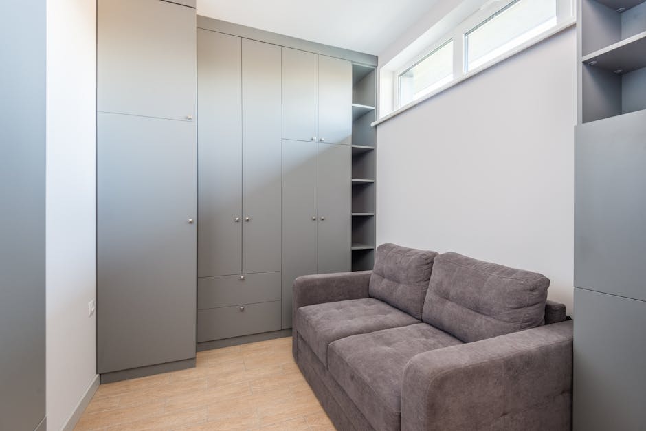 Interior view of a small room with a grey fabric sofa positioned against the right wall, adjacent to tall, grey storage cabinets with multiple doors and small round handles, and open shelving visible 
