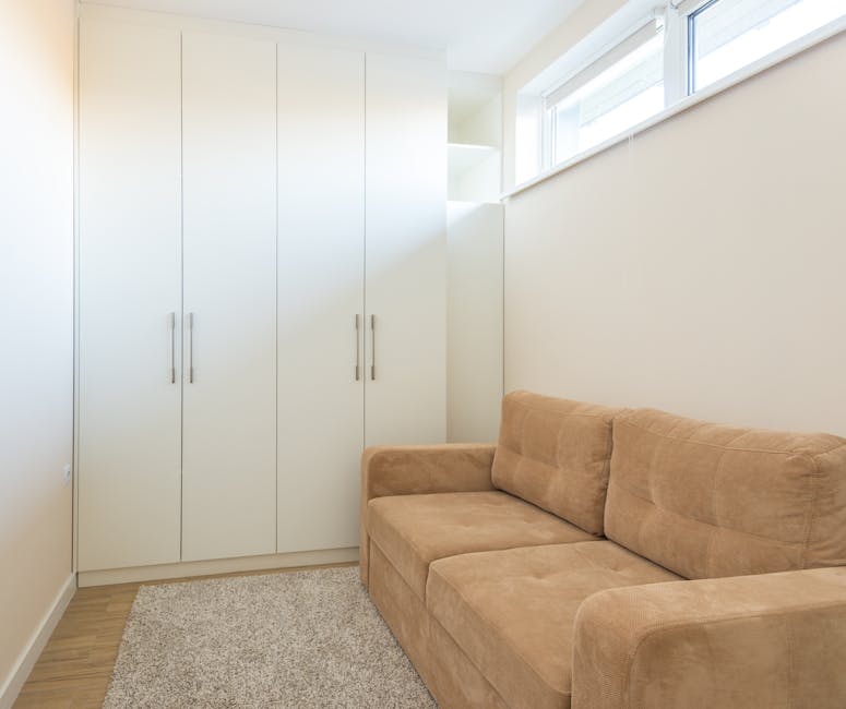 Interior view of a small room with a grey fabric sofa positioned against the right wall, adjacent to tall, grey storage cabinets with multiple doors and small round handles, and open shelving visible 