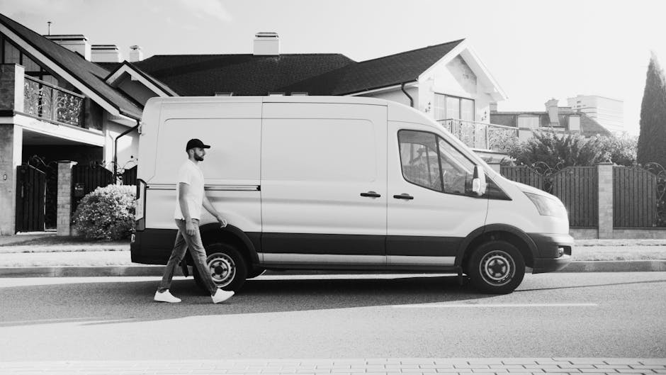 A man wearing a white T-shirt and jeans is standing on the pavement beside a white panel van, which has its rear doors open during a home relocation. He is placing a cardboard box onto a stack of simi