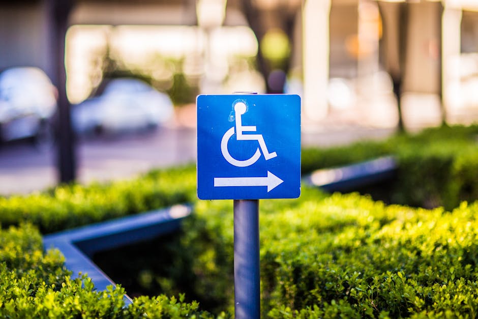 A blue accessible parking sign indicating wheelchair access with an arrow pointing to the right, mounted on a metal pole situated in a landscaped outdoor area with neatly trimmed green shrubs. In the background, blurred urban elements such as parked cars, pathways, and building structures are visible, suggesting the location is near a residential or public building. The sign is clearly visible and well-lit, with natural daylight highlighting the sign and surrounding greenery. This scene is relevant to house removals and moving logistics, as it depicts accessible parking guidance that may be utilized during local relocations, with Man With a Van Brockley's services supporting efficient furniture transport and packing in this environment.
