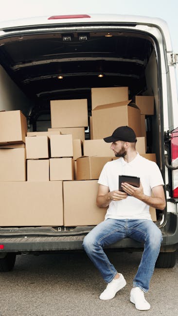 A man wearing a white t-shirt, jeans, and a black cap is sitting on the rear bumper of a white van with the back doors open during daytime. Inside the van, there are numerous cardboard boxes of varyin
