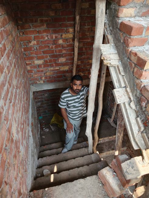 A man with dark hair and a beard, wearing a grey and white striped t-shirt and light grey trousers, stands on a set of rough, uneven brick and wooden stairs inside a narrow, unfinished brick-walled in